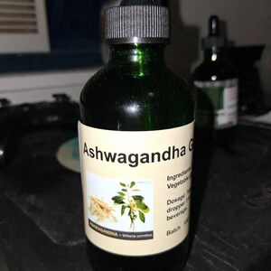 May include: A dark green glass bottle with a dropper top, labeled "Ashwagandha G". The label features text and an illustration of the ashwagandha plant. The bottle contains a dark liquid.