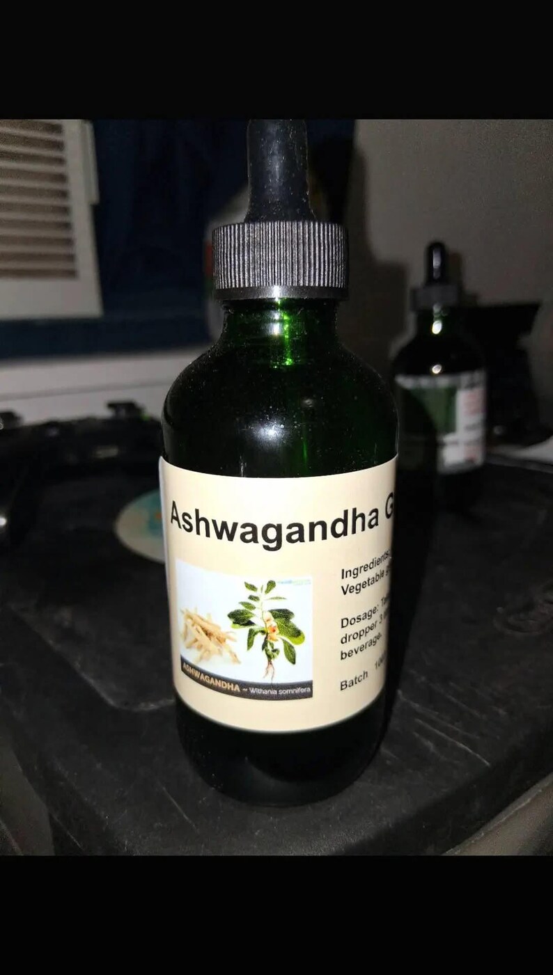 May include: A dark green glass bottle with a dropper top, labeled "Ashwagandha G". The label features text and an illustration of the ashwagandha plant. The bottle contains a dark liquid.