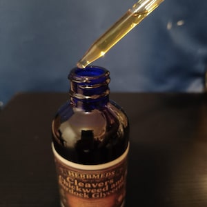 May include: A cobalt blue glass bottle with a dropper containing a golden liquid. The label reads HERBMEDS Cleavers Chickweed and Burdock Glycerite. The dropper is positioned above the open bottle neck, showing the amber-colored extract inside.