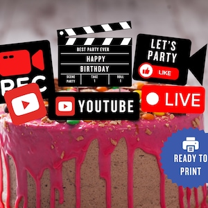 May include: A birthday cake with pink icing and colorful candies. The cake is decorated with black and red social media-themed toppers, including a clapperboard that reads "Happy Birthday." Other toppers include "REC," "YouTube," and "LIVE."