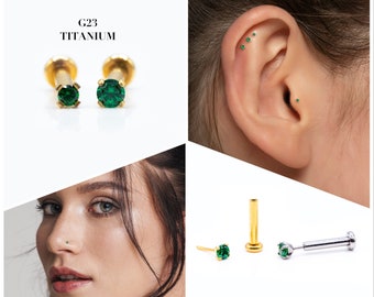 May include: Two gold titanium body jewelry studs with green gemstones. The studs are shown on a white background. The text "G23 TITANIUM" is shown above the studs.