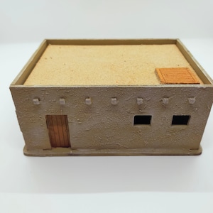 Op de afbeelding: Een klein, bruin, adobe-achtig gebouw met een zandkleurig dak. Het gebouw heeft een houten deur en twee ramen. Er is een klein houten luik op het dak.