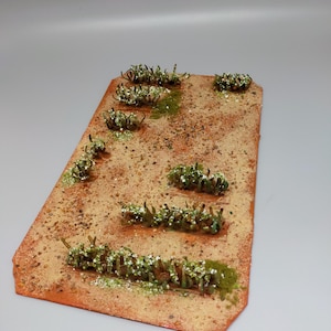 May include: A miniature desert terrain model with a sandy brown surface and green, leafy plants scattered across the surface. The model is made of a textured material and has a slightly irregular shape.