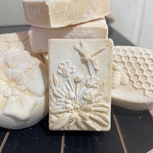 Puede incluir: Una colección de barras de jabón blancas con varios diseños en relieve, incluyendo una libélula, flores y un patrón de panal.