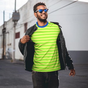 Può includere: Una persona indossa una giacca di pelle nera sopra una maglietta a righe verdi e gialle con colletto blu. La persona indossa anche occhiali da sole scuri e jeans scuri.