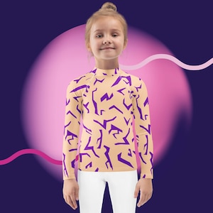 May include: A long-sleeved rash guard shirt with a mock neck. The shirt is tan with a repeating pattern of purple geometric shapes. The model is wearing white pants. The background is a gradient of purple and pink.