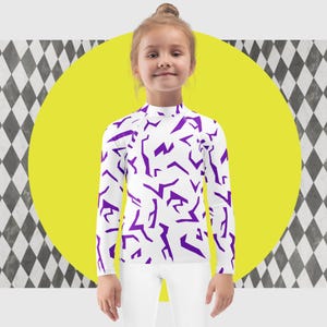 May include: A white long-sleeved shirt with a high collar, featuring a repeating pattern of purple geometric shapes. The shirt is worn by a child and paired with white pants. The background includes a yellow circle and a black and white diamond pattern.