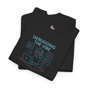Op de afbeelding: Zwart T-shirt met een blauwe en roze afbeelding van een persoon met een koptelefoon die codeert op een laptop. De tekst "Debugging the Vibe" staat op het T-shirt gedrukt.