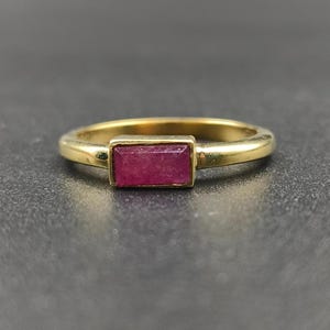 Puede incluir: Un anillo de oro con una gema rectangular de color rojo intenso. El anillo tiene una banda sencilla y un engaste de bisel que sujeta la gema de forma segura. El anillo se muestra sobre una superficie oscura y texturizada.