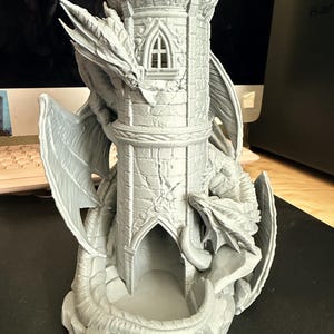 May include: A light gray 3D printed dice tower featuring a tall stone-textured turret with a pointed arch window and doorway. Two detailed dragons with large wings are wrapped around the exterior of the tower, one near the top and one at the base.