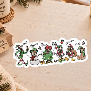 May include: A white sticker featuring embroidered-style illustrations of Goofy, Mickey Mouse as a snowman, Minnie Mouse as a Christmas tree, Daisy Duck, Donald Duck, and Pluto, all wearing festive holiday attire with their names written above them.