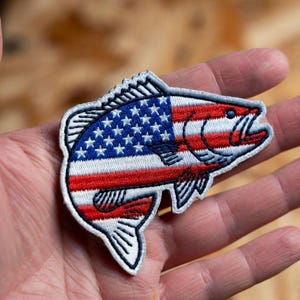 May include: An embroidered patch shaped like a fish, featuring the American flag pattern with red and white horizontal stripes and a blue section with white stars. The patch has a white border and is held in a palm, with the text Beverleyharveyst visible.