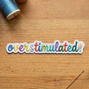 May include: An embroidered patch featuring the word overstimulated in colourful, lowercase letters with a small flower at the end. The patch is surrounded by blue and purple thread spools, a metal thimble, and a sewing needle on a wooden surface.