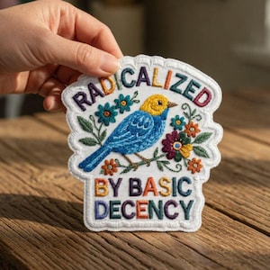 Puede incluir: Parche bordado con un pájaro azul y cabeza amarilla, rodeado de flores coloridas y el texto "RADICALIZED BY BASIC DECENCY". El parche tiene un borde festoneado blanco.