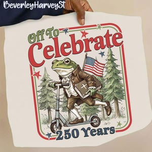 Könnte beinhalten: Cremefarbenes T-Shirt mit einem Cartoon-Frosch auf einem Roller, der ein Buch und eine amerikanische Flagge hält. Der Frosch trägt Kleidung aus der Kolonialzeit. Der Text lautet "Off To Celebrate 250 Years" in Rot und Grün.