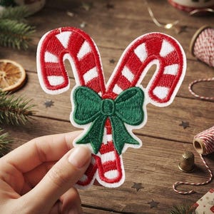 May include: An embroidered patch featuring two crossed candy canes with red and white diagonal stripes, accented by a central dark green bow. The patch has a white border and a textured thread finish, depicting a classic holiday motif.