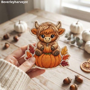 Puede incluir: Una pegatina troquelada que muestra una vaca Highland marrón y esponjosa sentada sobre una calabaza naranja grande, sosteniendo una calabaza más pequeña. El diseño incluye hojas de otoño coloridas y el texto Beverleyharveyst.