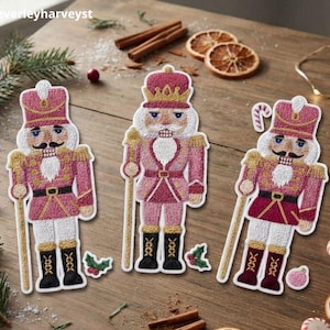 Könnte beinhalten: Drei gestickte Nussknacker-Aufnäher in Rosa-, Gold- und Weißtönen. Jede Figur trägt eine hohe Mütze, eine Uniform mit goldenen Akzenten und schwarze Stiefel. Ein Nussknacker hält einen Stab, ein anderer eine Zuckerstange, alle mit feinen Stickereien.
