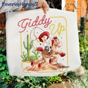 May include: White tote bag with a cartoon illustration of a cowgirl riding a horse, with the words "Giddy Up" in red and yellow. The design includes cacti and a rope, evoking a Western theme.