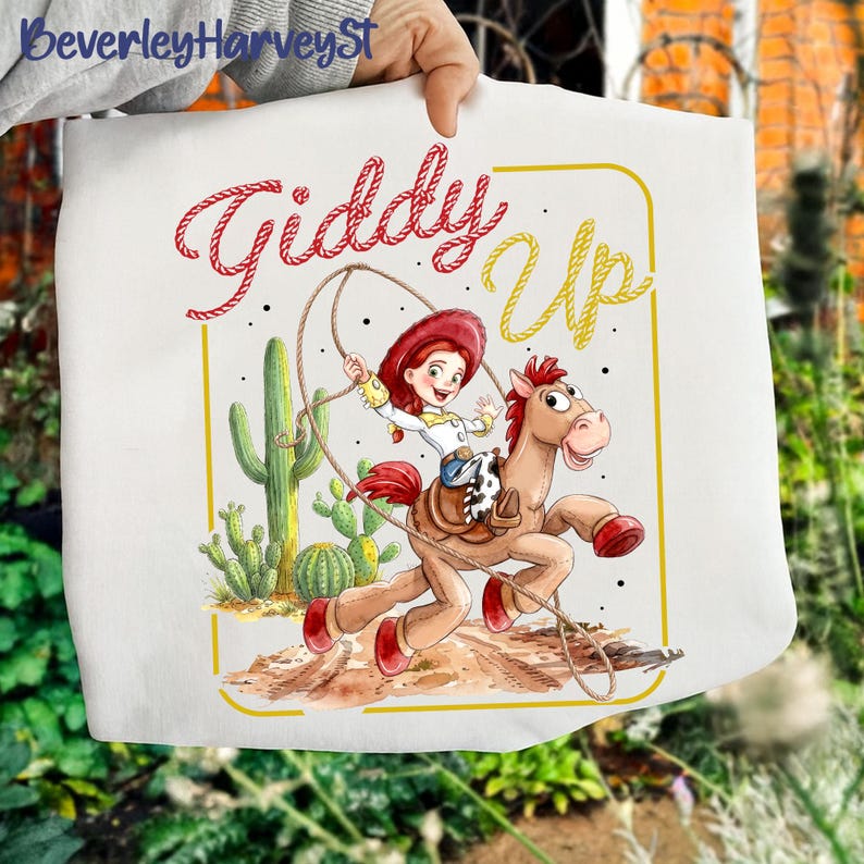 May include: White tote bag with a cartoon illustration of a cowgirl riding a horse, with the words "Giddy Up" in red and yellow. The design includes cacti and a rope, evoking a Western theme.
