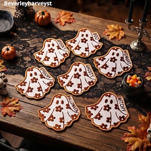 Puede incluir: Seis posavasos blancos con forma de fantasma, con patrones florales marrones y bordes marrones. Cada posavasos presenta una palabra única impresa en marrón: SPOOKY, BOO, MIDNIGHT, SPOOCHY, GHOSTY, HALLOWEEN y WITCHY sobre una superficie.