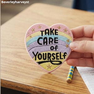 Könnte beinhalten: Ein herzförmiger Stickaufnäher mit pastellfarbenem Regenbogenhintergrund, goldenen Sternen und dem schwarzen Text "TAKE CARE of YOURSELF" in der Mitte. Der Aufnäher hat einen rosa gestickten Rand und wird über einer Holzoberfläche gehalten.