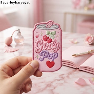 May include: A pink embroidered patch shaped like a soda can featuring the text Girly Pop in pink and purple script. The design includes a pair of red cherries with a light green bow, a red heart, and small star accents on a light pink background.