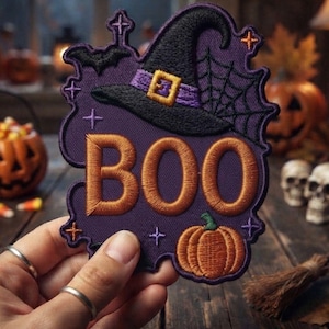 May include: A purple embroidered patch featuring the text "BOO" in orange, a black witch hat with a purple band and yellow buckle, a spider web, a black bat, a small orange pumpkin, and several small star-shaped accents in orange and purple.