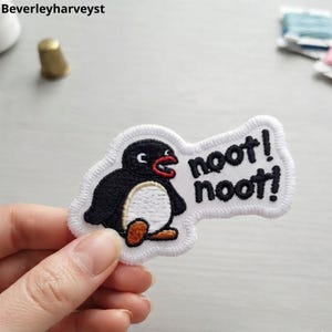 Op de afbeelding: Een geborduurde strijkpatch met een zwart-witte pinguïn met een oranje snavel en voeten. De pinguïn is afgebeeld met een open mond en de tekst "noot! noot!" is in zwart garen op een witte achtergrond geborduurd.