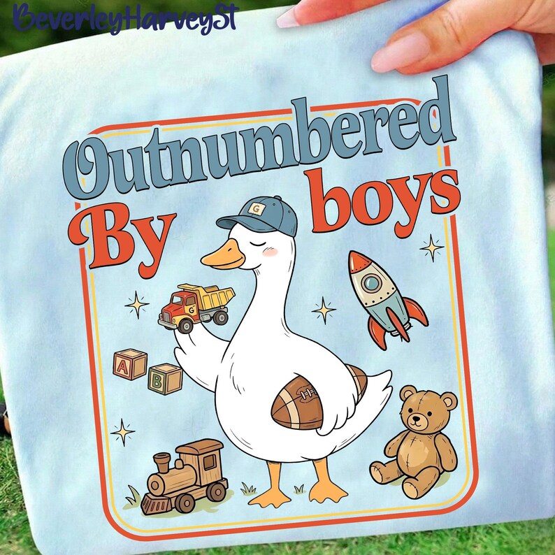 May include: Pale blue t-shirt featuring a cartoon goose in a cap, holding a football, toy truck, and alphabet blocks. The text "Outnumbered By Boys" is in a retro style, with a rocket, train, teddy bear, and stars.