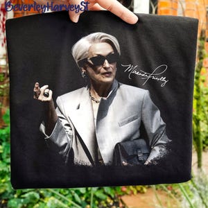 Può includere: Borsa tote nera con un ritratto di una donna con occhiali da sole e un abito grigio. L'immagine include la firma "Miranda Priestly" in grafia bianca. La borsa è tenuta da una mano.