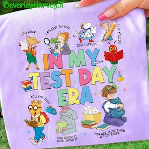 May include: A lilac t-shirt with the words "IN MY TEST DAY ERA" in bright letters. The design includes cartoon characters like a llama, mouse, and elephant, along with the phrases "YOU ROCK", "I BELIEVE IN YOU", and "YOU GOT THIS". The shirt is decorated with books and test papers.