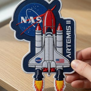 May include: Embroidered patch featuring a space shuttle design with the NASA logo and the text "ARTEMIS II." The patch has a blue background with white, red, and yellow accents. The patch is held by a hand.