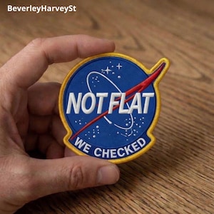 Puede incluir: Un parche circular con fondo azul, borde amarillo y el texto "NOT FLAT WE CHECKED" en blanco. El parche presenta un diseño gráfico rojo y blanco, que se asemeja a la trayectoria de un cohete. El parche está sostenido por una mano.