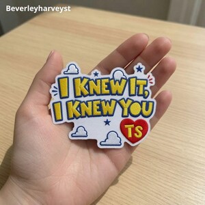 Puede incluir: Un parche bordado blanco con borde en forma de nube que presenta el texto amarillo I KNEW IT, I KNEW YOU en fuente negrita. El diseño incluye pequeñas estrellas azules, contornos de nubes blancas y un corazón rojo con las letras TS.