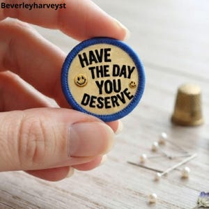 Puede incluir: Un parche bordado circular con un centro de color crema y un borde azul. El texto "HAVE THE DAY YOU DESERVE" está bordado en negro, acompañado de dos pequeños iconos de cara sonriente en tono dorado a la izquierda y a la derecha.