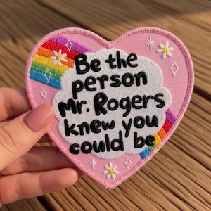 May include: A pink heart-shaped embroidered patch with the text "Be the person Mr. Rogers knew you could be." The patch features a rainbow, white cloud, and small floral and diamond accents. The patch is a decorative item.