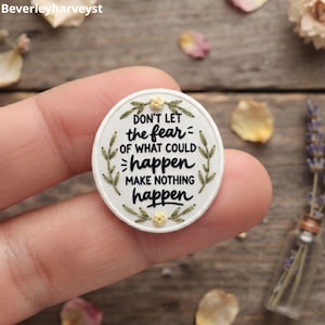 Può includere: Una toppa ricamata circolare bianca con il testo nero "Don't let the fear of what could happen make nothing happen". Il disegno è incorniciato da foglie ricamate verdi e piccoli fiori gialli sui bordi superiore e inferiore.