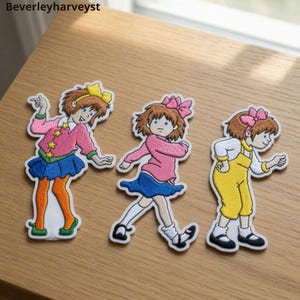 Può includere: Tre toppe ricamate raffiguranti un personaggio dei cartoni animati con capelli castani. La prima indossa un cardigan rosa con stelle gialle, una gonna blu e collant arancioni. La seconda indossa un maglione rosa e una gonna blu. La terza indossa una salopette gialla.