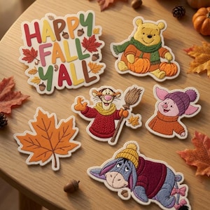 Puede incluir: Una colección de parches bordados con el texto HAPPY FALL Y'ALL, Winnie the Pooh con calabazas, Tigger con una escoba, Piglet con gorro, Eeyore con suéter y una hoja de arce naranja, dispuestos sobre una superficie de madera.