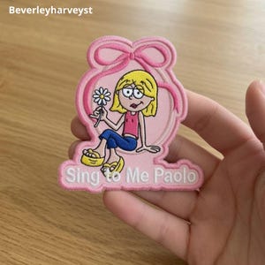 Op de afbeelding: Een roze geborduurde patch met een stripfiguur met blond haar, een roze topje en een blauwe broek, die een witte margriet vasthoudt. Het figuur is omlijst door een roze lint. De tekst Sing to Me Paolo is onderaan in het wit geborduurd.