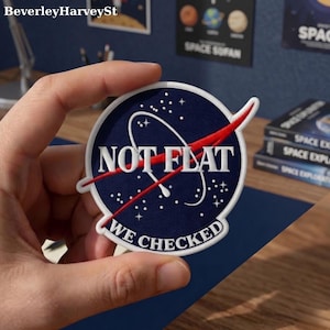 Puede incluir: Un parche azul marino con texto blanco que dice "NOT FLAT" y "WE CHECKED". El parche presenta un trazo rojo y estrellas blancas, que se asemejan a un logotipo de la NASA. El parche está sostenido por una mano.