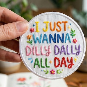 May include: A circular white fabric patch featuring colorful hand-embroidered text that reads I JUST WANNA DILLY DALLY ALL DAY. The lettering uses various thread colors including yellow, blue, pink, purple, green, and red, accented by small floral motifs.