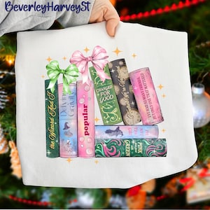 May include: White sweatshirt featuring a stack of colorful books with titles like "Popular" and "Changed for Good." The books are adorned with bows and surrounded by star accents. The text "BeverleyHarveySt" is at the top.