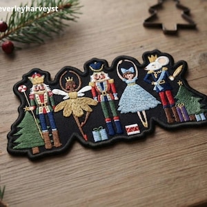 May include: A black embroidered patch featuring Nutcracker-themed characters including two nutcracker soldiers, a fairy, a ballerina, and a mouse king. The design includes a Christmas tree, wrapped gifts, and a drum, all outlined in black thread.