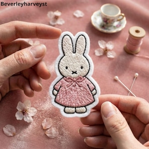 Pode incluir: Um emblema bordado apresentando uma personagem de coelho branco com orelhas compridas, olhos pretos e uma boca em forma de cruz preta, vestindo um vestido rosa com um laço no decote. O emblema tem uma borda de feltro branco.