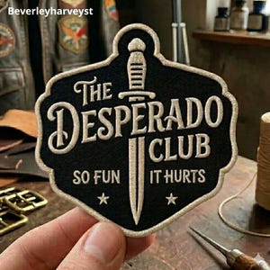 Könnte beinhalten: Ein schwarzer bestickter Aufnäher mit cremefarbenem Rand und einem zentralen Dolchmotiv. Der Text lautet The Desperado Club und So Fun It Hurts, ergänzt durch zwei kleine Sterne. Der Aufnäher hat eine schildartige Form mit gewelltem Rand.
