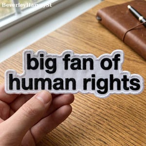 May include: White embroidered patch with the text "big fan of human rights" in black, with a white border. The patch is held in a hand, with a wooden surface in the background.