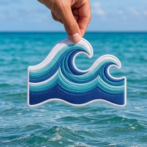 May include: Embroidered wave patch in shades of blue and white, held against a backdrop of the ocean and sky. The patch features a detailed design with a white border and the text "BeverleyHarveySt" in the top left corner.