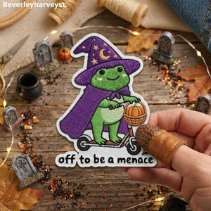 May include: An embroidered patch featuring a green frog wearing a purple witch hat with a yellow moon and stars, and a purple cape. The frog is riding a scooter and holding a basket with a pumpkin. The text reads, off to be a menace.
